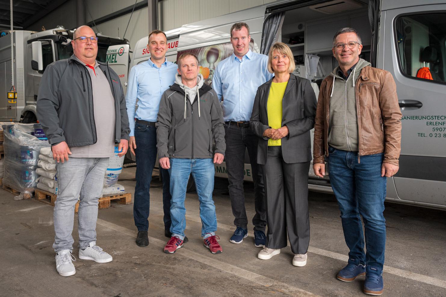 Sechs Personen stehen lächelnd in einer Lagerhalle, Hintergrund zeigt Nutzfahrzeuge und Stapel mit Materialien.