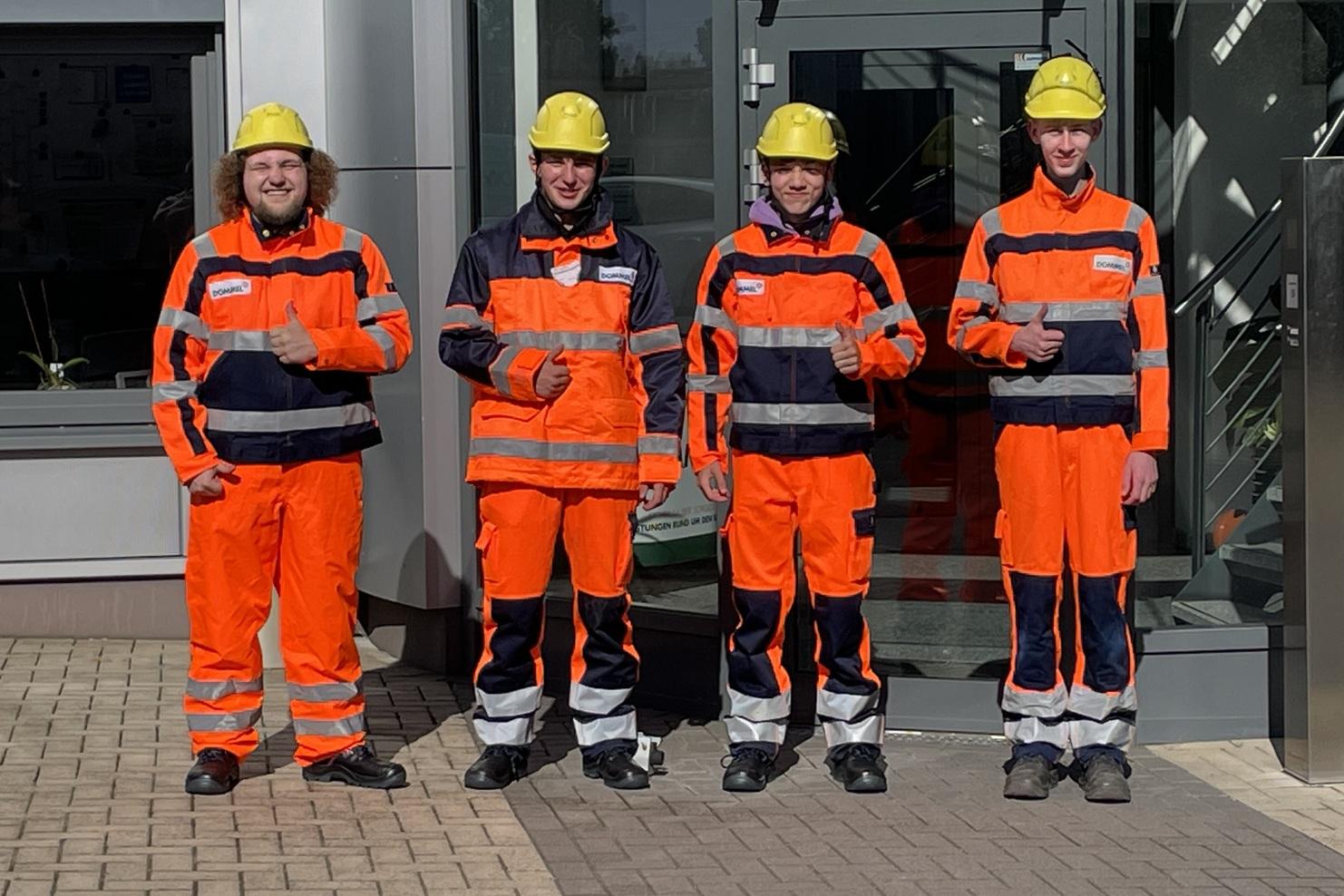 Vier Personen in orangefarbener Arbeitskleidung und Helmen stehen lächelnd vor einem Gebäude.