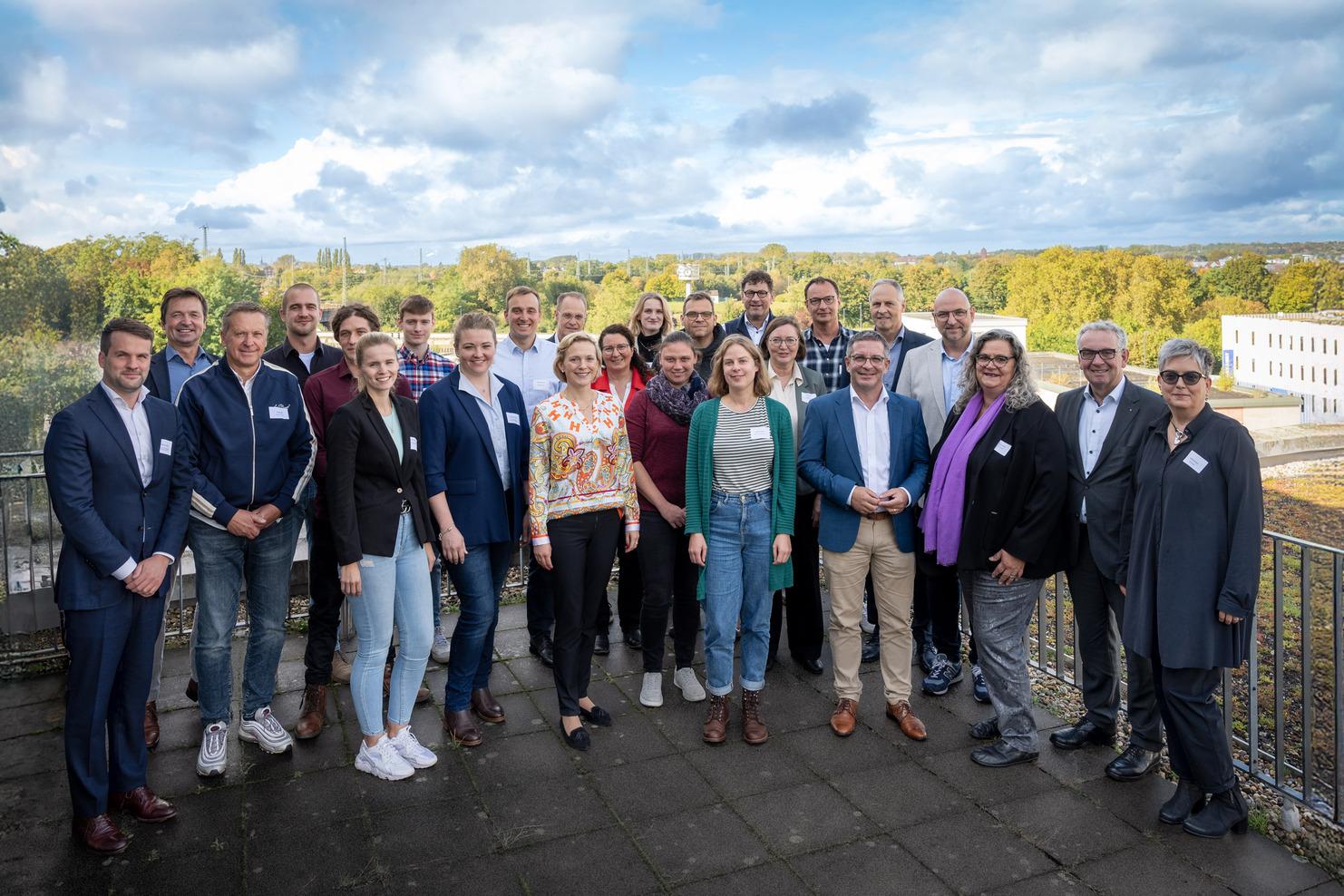 Gruppenfoto von 24 Personen in businessgerechter Kleidung, stehend auf einer Terrasse mit grünem Hintergrund und bewölktem Himmel.