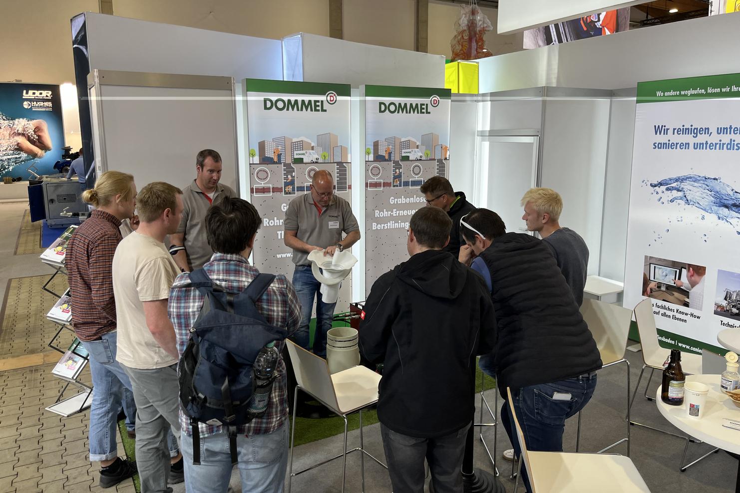 Gruppe von Menschen am Messestand von DOMMEL, die sich über Rohrsanierungsdienstleistungen informieren.