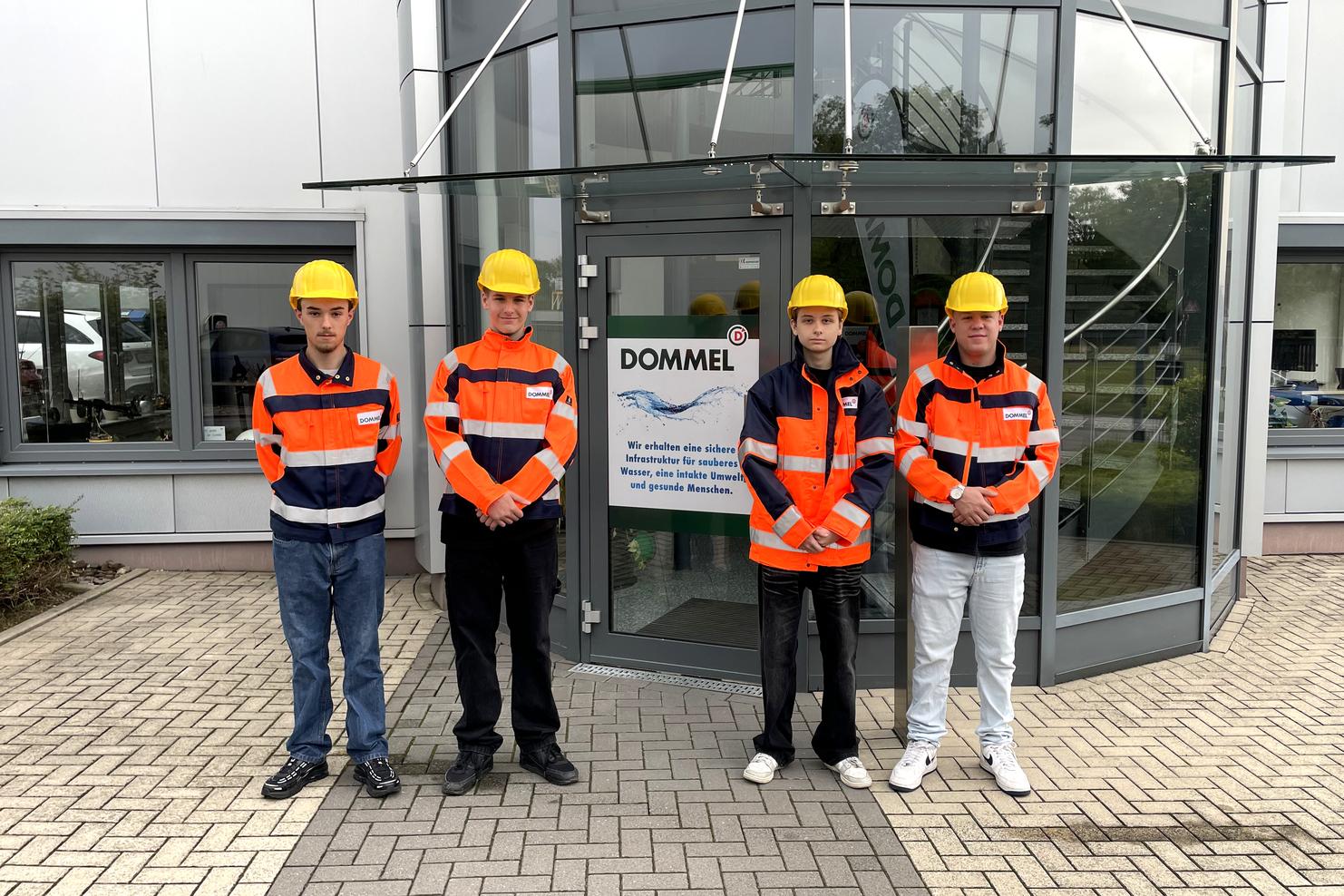 Vier Personen in Warnkleidung und Helmen stehen vor einem Firmengebäude mit dem Logo „DOMMEL“.