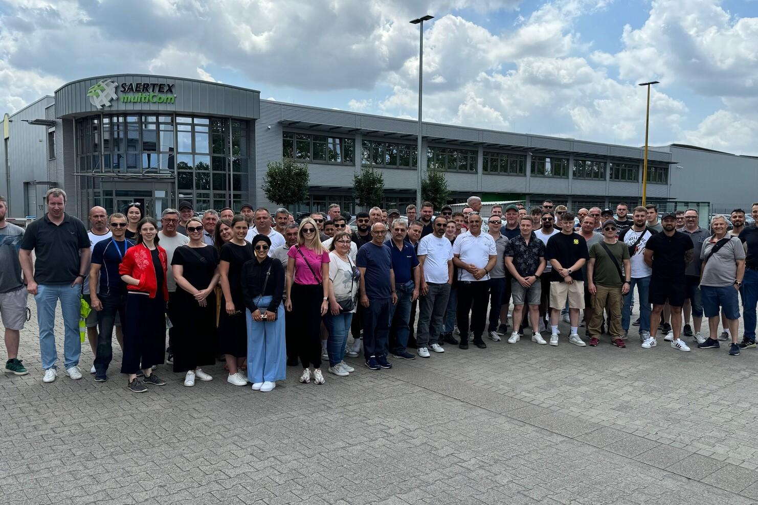 Gruppenfoto von über 50 Personen vor dem Eingang des Saertex-Werks unter einem bewölkten Himmel.