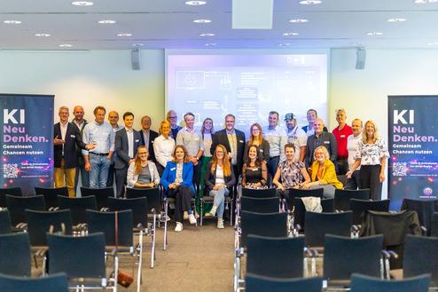 Gruppenfoto von Teilnehmern einer Konferenz zu Künstlicher Intelligenz, mit Bannern im Hintergrund und Präsentation an der Wand.
