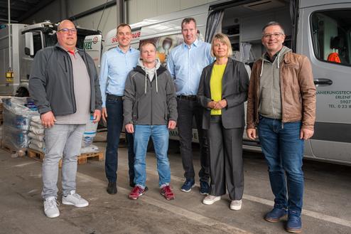 Sechs Personen stehen lächelnd in einer Lagerhalle, Hintergrund zeigt Nutzfahrzeuge und Stapel mit Materialien.