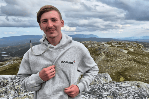Junger Mann in grauem Hoodie mit Aufdruck steht auf einem Berg mit gebirgiger Landschaft im Hintergrund.