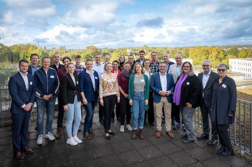 Gruppenfoto von 24 Personen in businessgerechter Kleidung, stehend auf einer Terrasse mit grünem Hintergrund und bewölktem Himmel.
