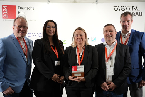 Gruppenschau von fünf Personen, die den Deutschen Bau Preis 2024 halten, vor einem beschrifteten Hintergrund.