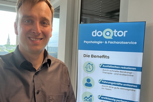 Mann im Büro lächelt vor einem Banner des Unternehmens doQtor, das psychologische Dienstleistungen bewirbt.