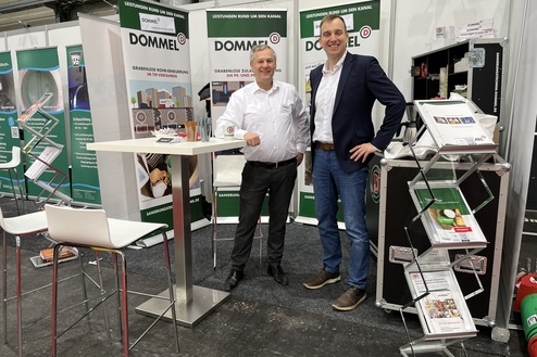 Zwei Männer stehen lächelnd an einem Messestand mit Dommel-Branding und Informationsmaterialien in einer Messehalle.