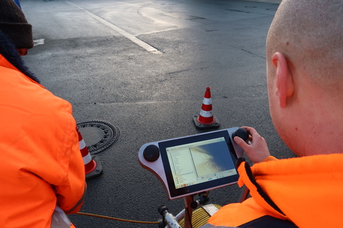 Zwei Arbeiter in orangefarbener Kleidung bedienen einen Monitor zur Inspektion einer Straßenoberfläche.