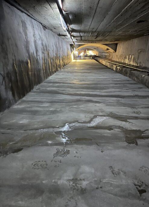 Blick entlang eines unterirdischen Ganges mit glänzender, nasser Betonoberfläche und schwachem Licht.