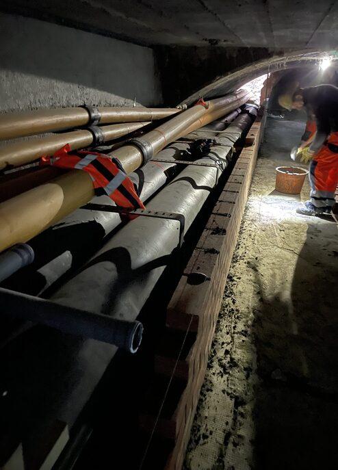 Überblick über eine dunkle Baustelle mit Rohrleitungen und einem Arbeiter, der mit einem Eimer auf dem Boden arbeitet.