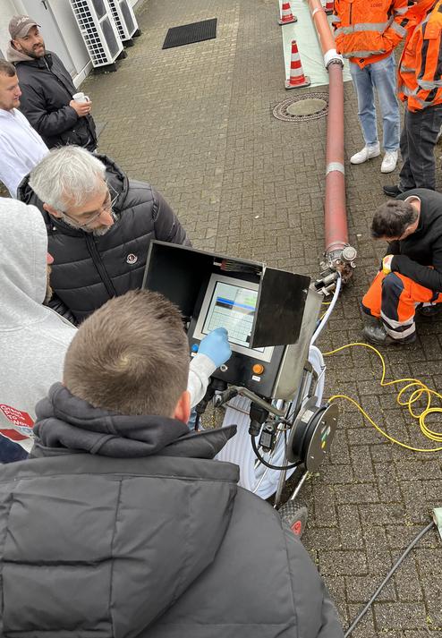 Gruppe von Fachleuten betrachtet ein Gerät mit Bildschirm und Bedienelementen vor einer Rohrleitung im Freien.