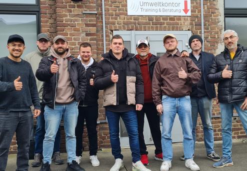 Gruppe von acht Männern, die vor einem Gebäude mit einem Schild für ein Beratungsunternehmen stehen und Daumen hoch zeigen.