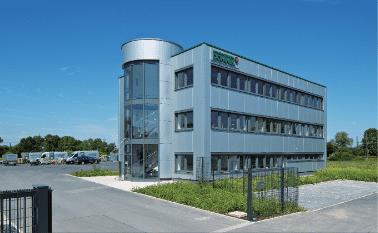 Moderne, mehrstöckige Bürogebäude mit Glasfassade und grüner Dachkante, umgeben von Parkplätzen und Wiese.