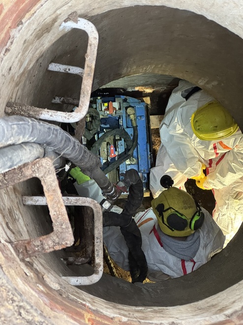 Arbeiter in Schutzausrüstung reparieren Geräte in einem engen, runden Schacht mit Leitungen und Werkzeugen.