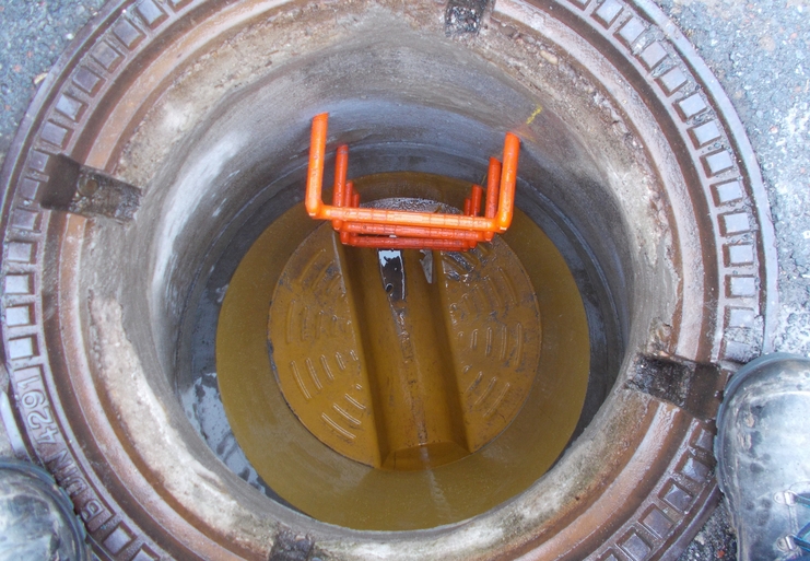 Blick in einen Schacht mit metallischem Boden und orangefarbener Leiter, umgeben von einem runden Gitterdeckel.