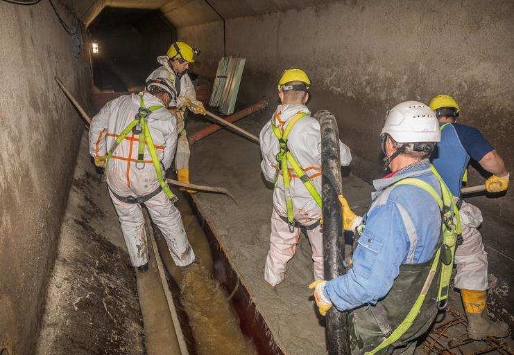 Fünf Arbeiter in Schutzkleidung mischen Beton in einem unterirdischen Kanal mit Wasserablauf und schwachem Licht.