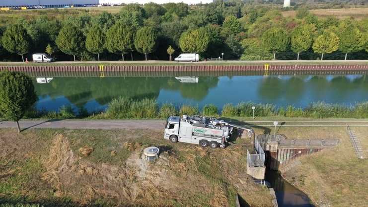 Lkw mit Abwassersammeltechnik am Ufer eines Kanals, umgeben von Bäumen und reflektierendem Wasser.