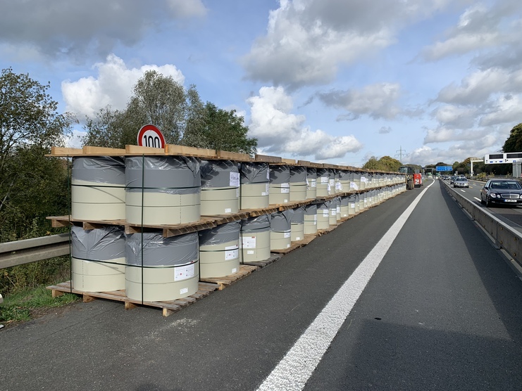 Reihe von großen, verpackten Fässern auf Holzpaletten entlang einer Autobahn mit Verkehr und Geschwindigkeitsbegrenzungsschild.