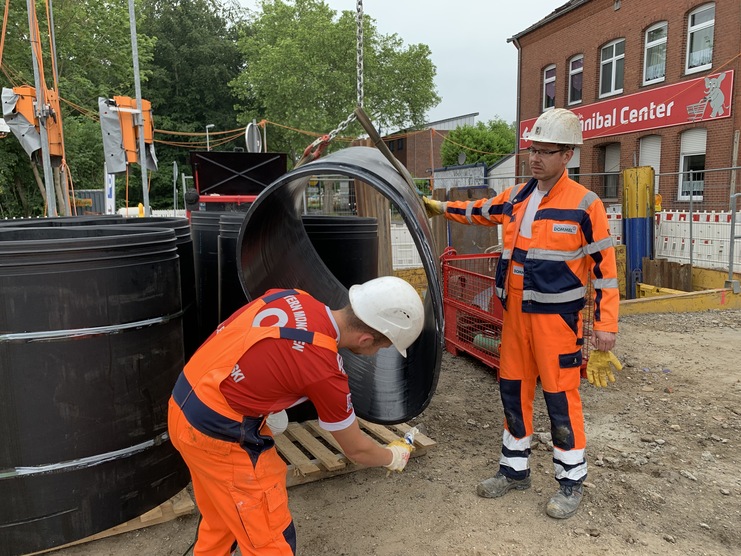 Zwei Bauarbeiter in Schutzkleidung arbeiten mit großen schwarzen Rohrteilen auf einer Baustelle.
