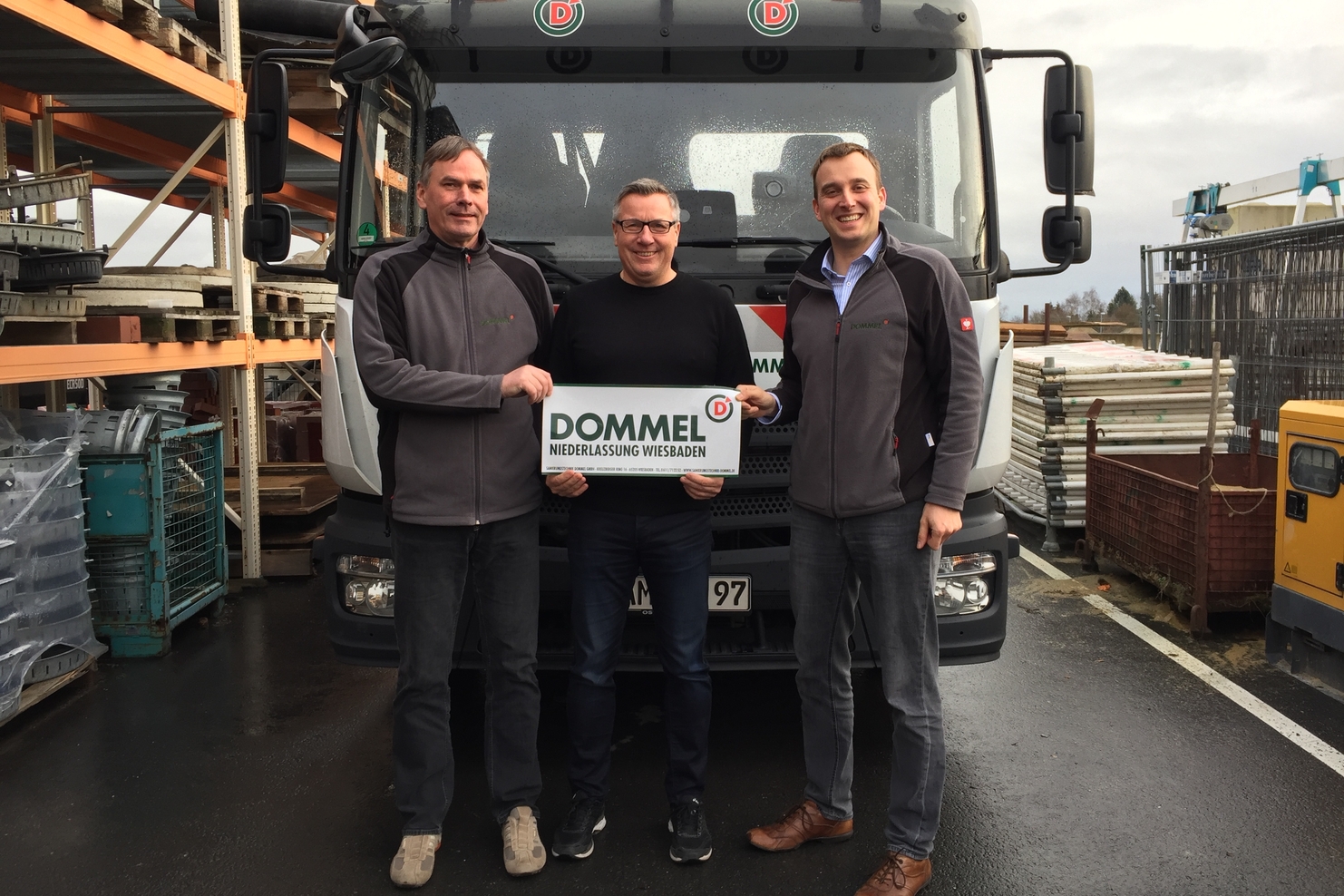 Drei Männer stehen vor einem Lkw und halten ein Schild mit der Aufschrift „DOMMEL NIEDERLASSUNG WIESBADEN“.