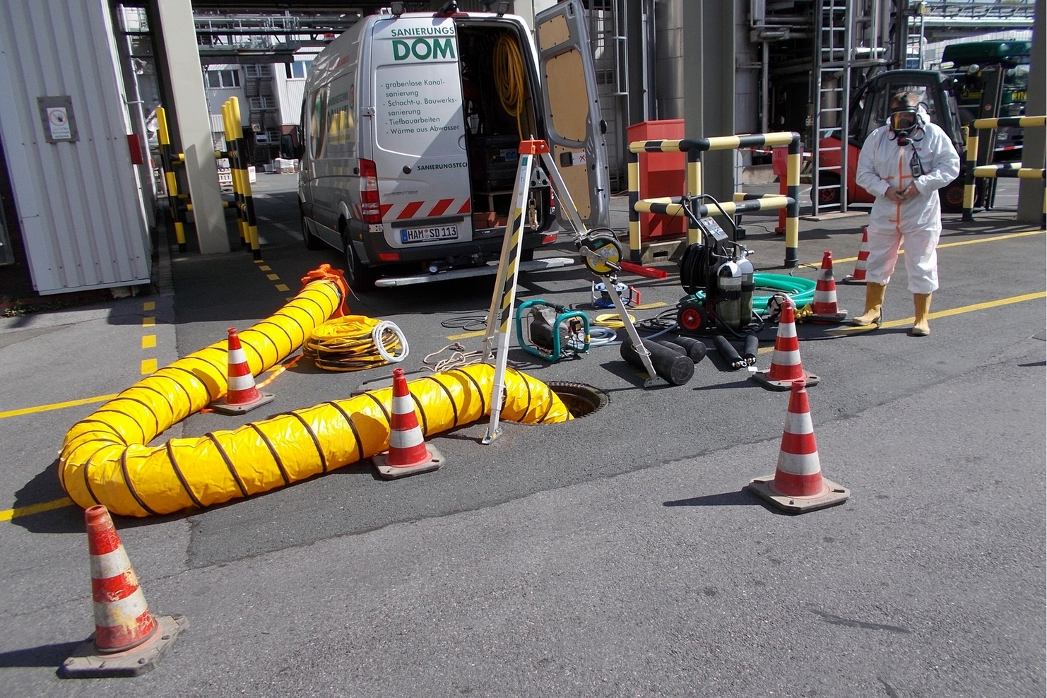 Sanierungsteam in Schutzkleidung arbeitet neben einem offenen Schacht, umgeben von Warnbaken und Arbeitsger&auml;ten.