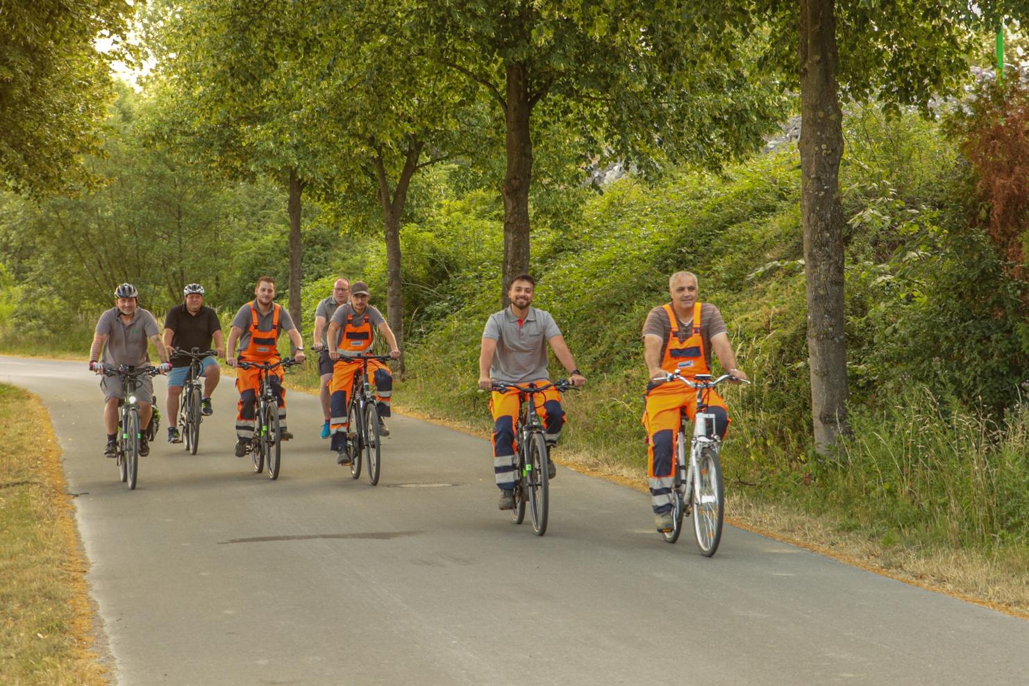 Sechs Männer in Arbeitskleidung radeln auf einem gepflasterten Weg unter Bäumen.