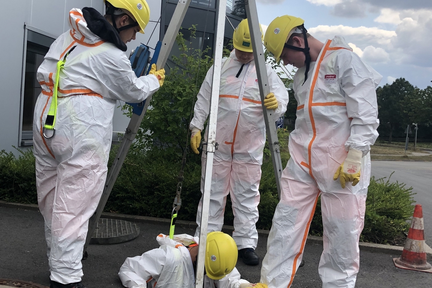 Vier Personen in Schutzanzügen und Helmen arbeiten an einem Schacht, eine Person ist teilweise darin.