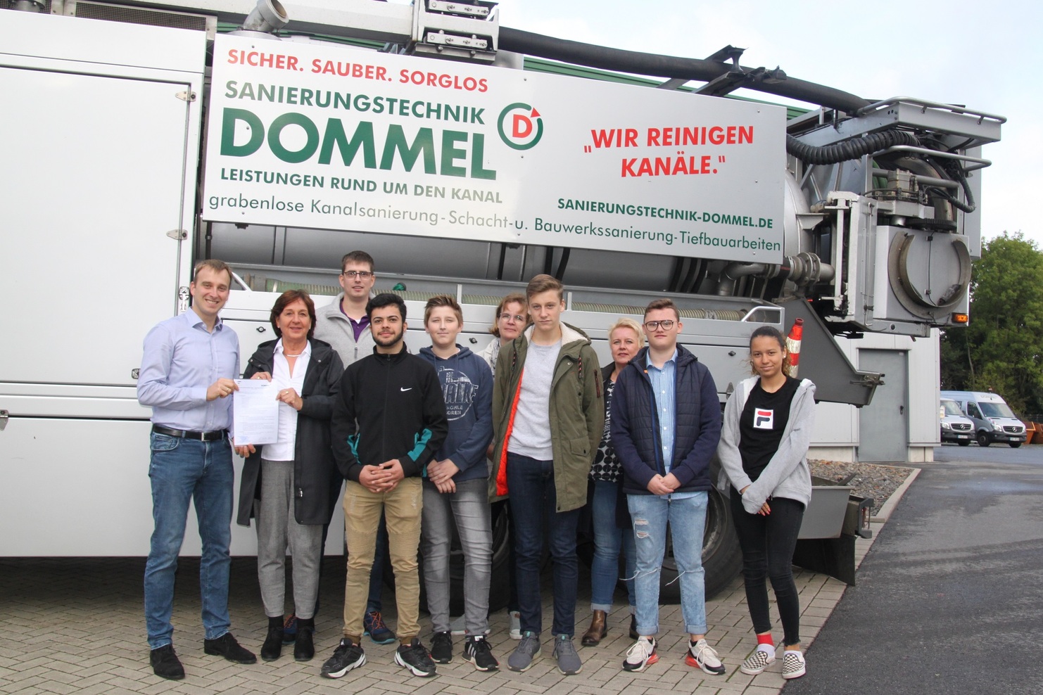 Gruppe von Personen steht vor einem Fahrzeug mit dem Schriftzug „Sanierungstechnik Dommel“ und Informationen zu den Dienstleistungen.