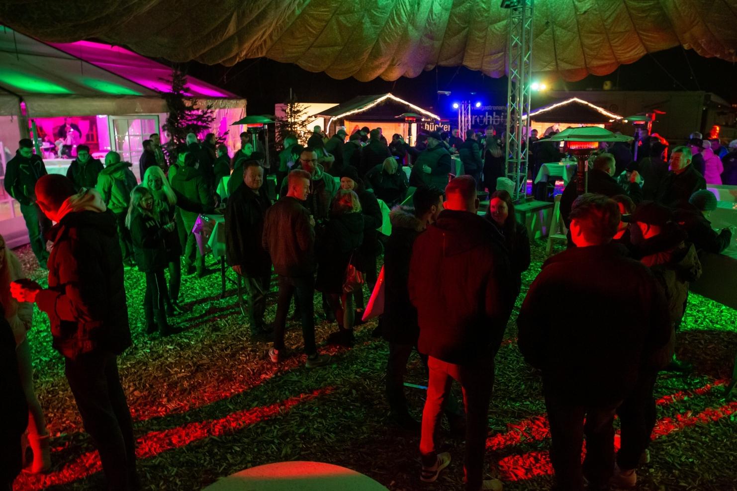 Menschenmenge bei Nacht auf einer festlich beleuchteten Veranstaltung unter einem Zelt mit grünen und roten Lichtern.
