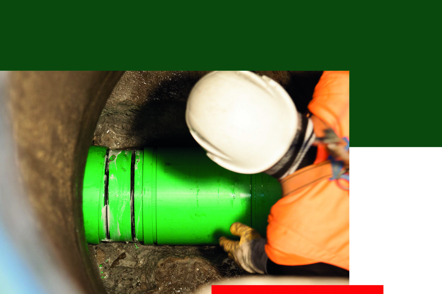 Nahaufnahme eines Arbeiters mit Helm, der eine grüne Rohrleitung im Erdreich befestigt. Hintergrund ist dunkel und schmutzig.