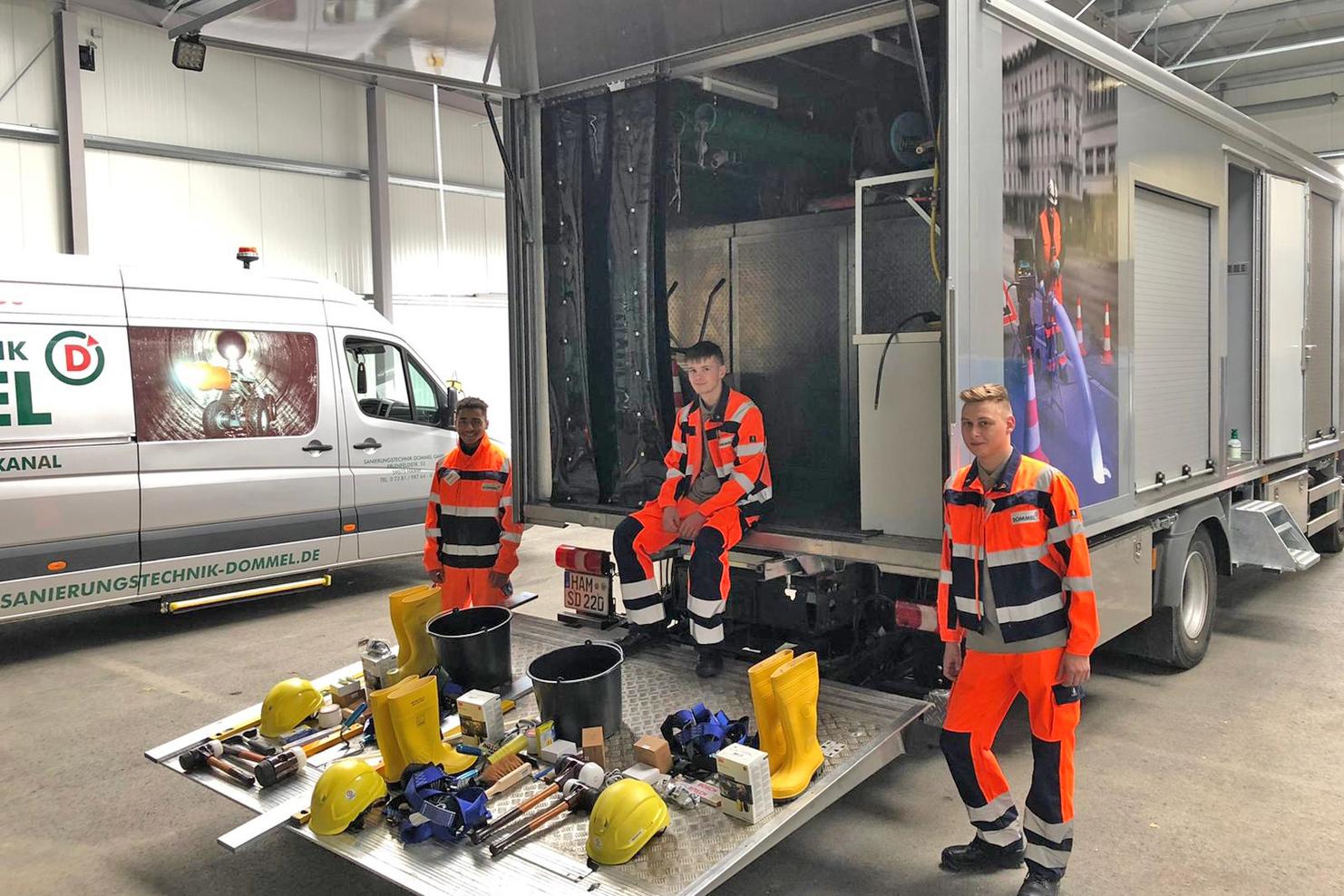 Zwei Männer in orangen Arbeitsanzügen stehen neben einem Lkw, umgeben von Werkzeugen und Ausrüstung auf einer Ladefläche.