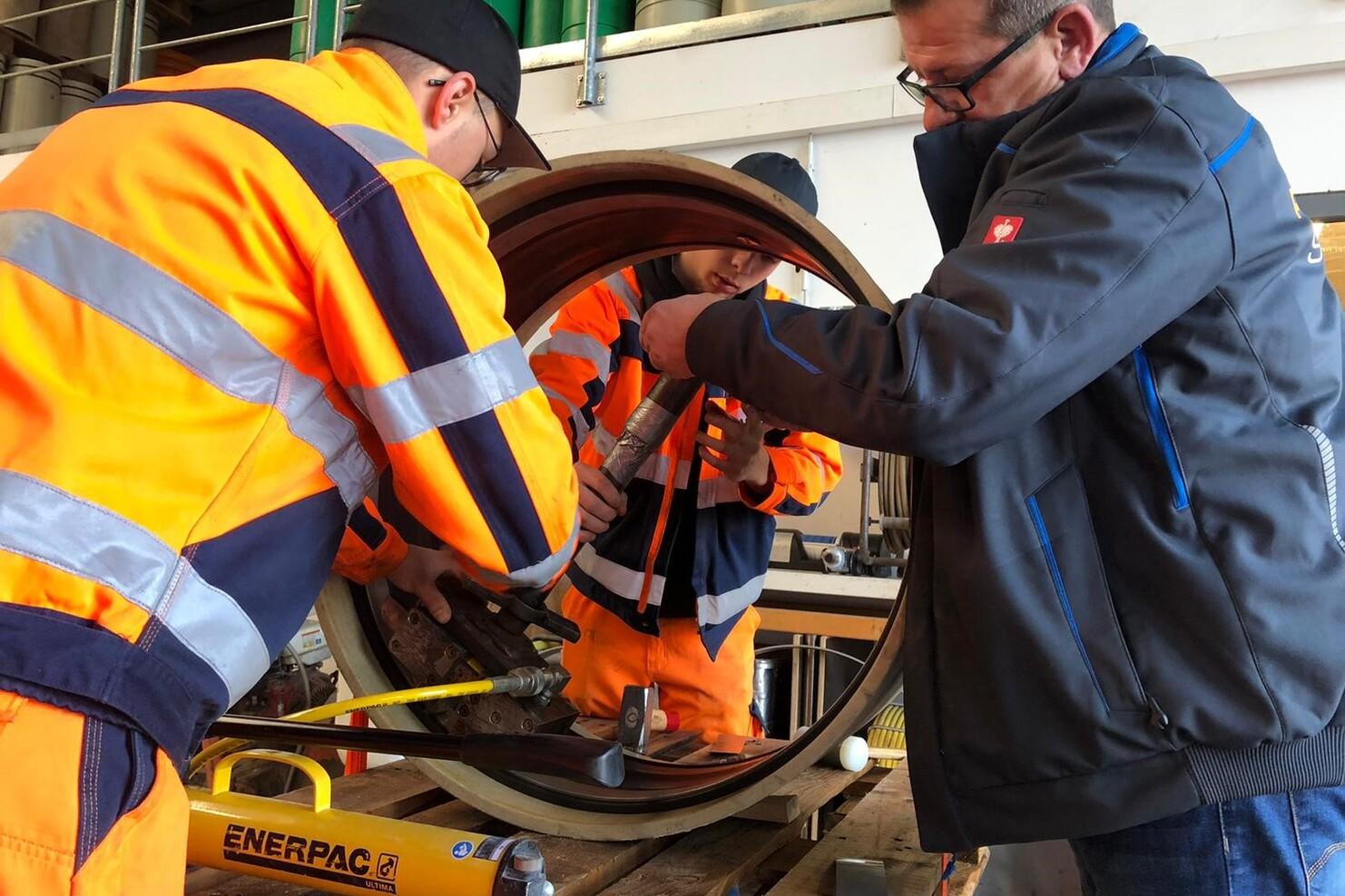 Drei Personen in Warnkleidung arbeiten an einem großen zylindrischen Objekt auf einer Werkbank.