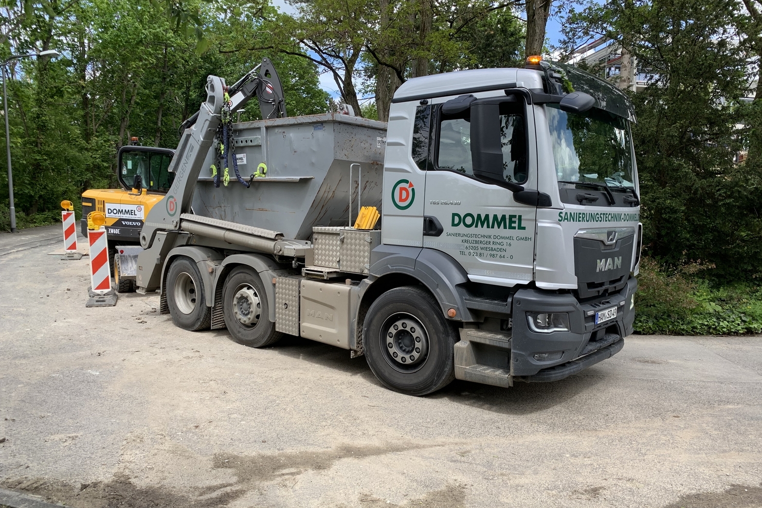 Lkw der Firma Dommel transportiert einen Abfallcontainer auf einer Baustelle, umgeben von Baumal und Verkehrszeichen.