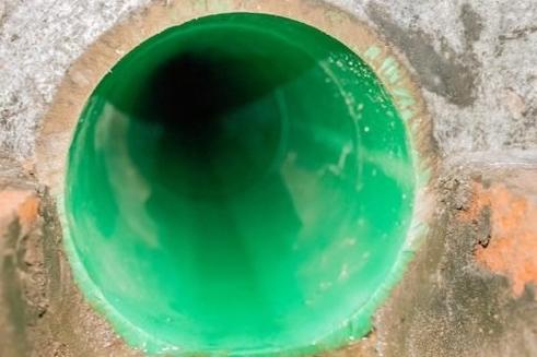 Nahaufnahme eines grünen Abflussrohrs, das in eine dunkle Öffnung führt, mit sichtbarem Mauerwerk außen.