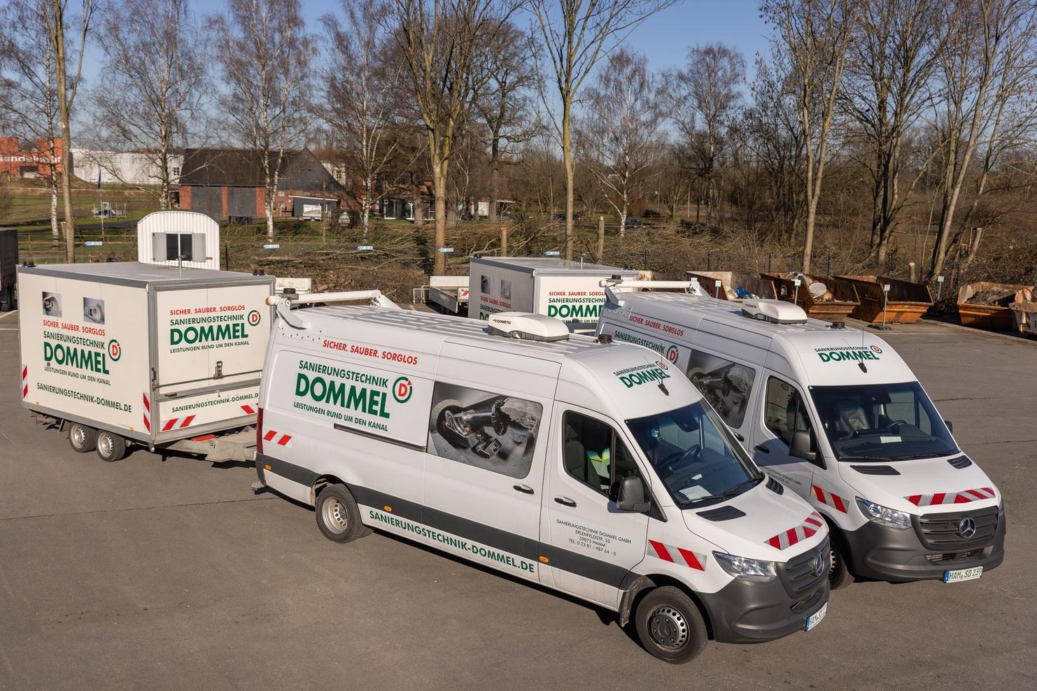 Drei Firmenfahrzeuge von Dommel mit Anhänger stehen auf einem Gelände, umgeben von Bäumen und Bauabbrüchen im Hintergrund.