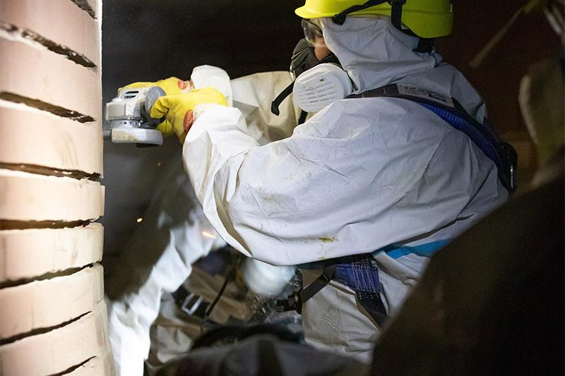 Arbeiter in Schutzkleidung verwenden ein Werkzeug, um an einem ziegelverkleideten Wandabschnitt zu arbeiten.