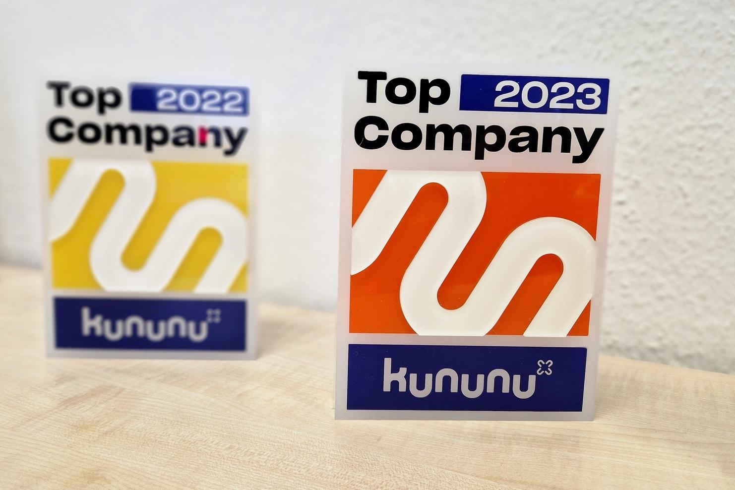 Zwei Auszeichnungen von Kununu für "Top Company 2022" und "Top Company 2023" auf einem Tisch.