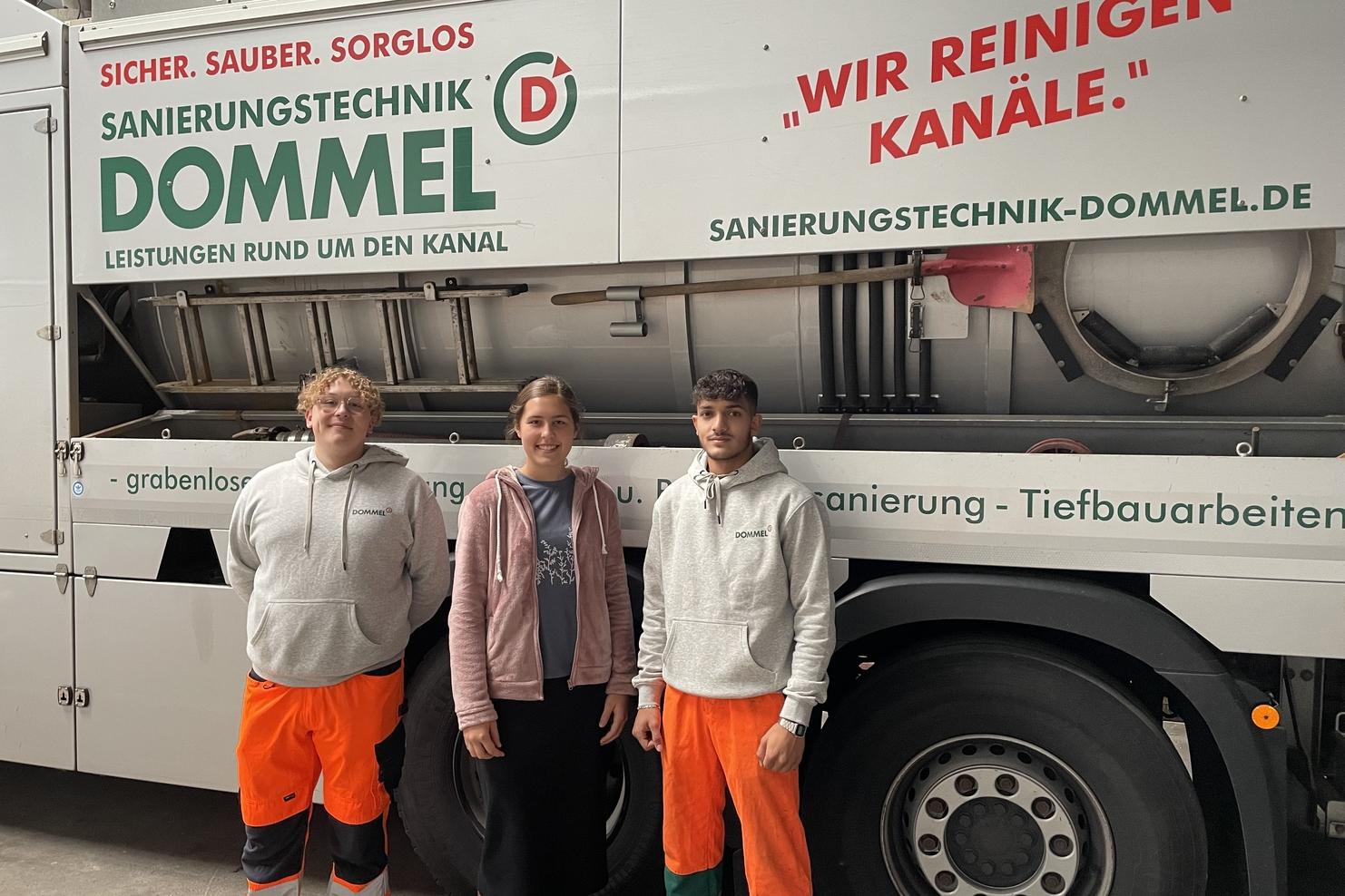 Drei Personen stehen vor einem Fahrzeug der Firma Dommel mit einem großen Werbeslogan zur Kanalsanierung.