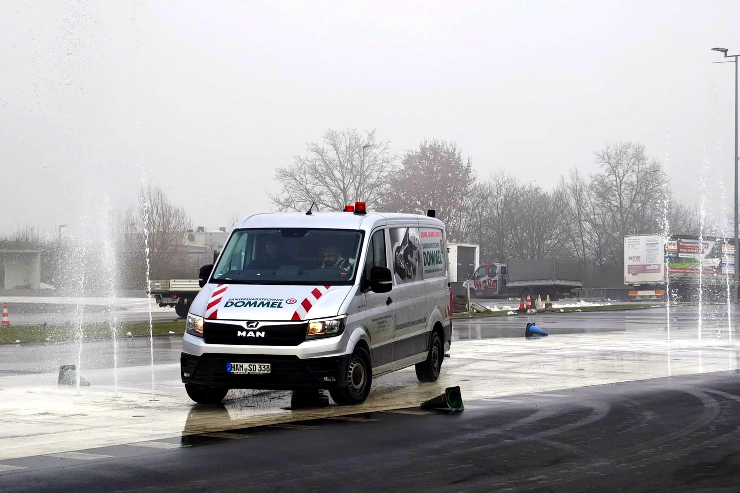 Ein MAN-Transporter fährt auf einer feuchten Verkehrsübungsfläche, während Spritzwasserfontänen aktiv sind.