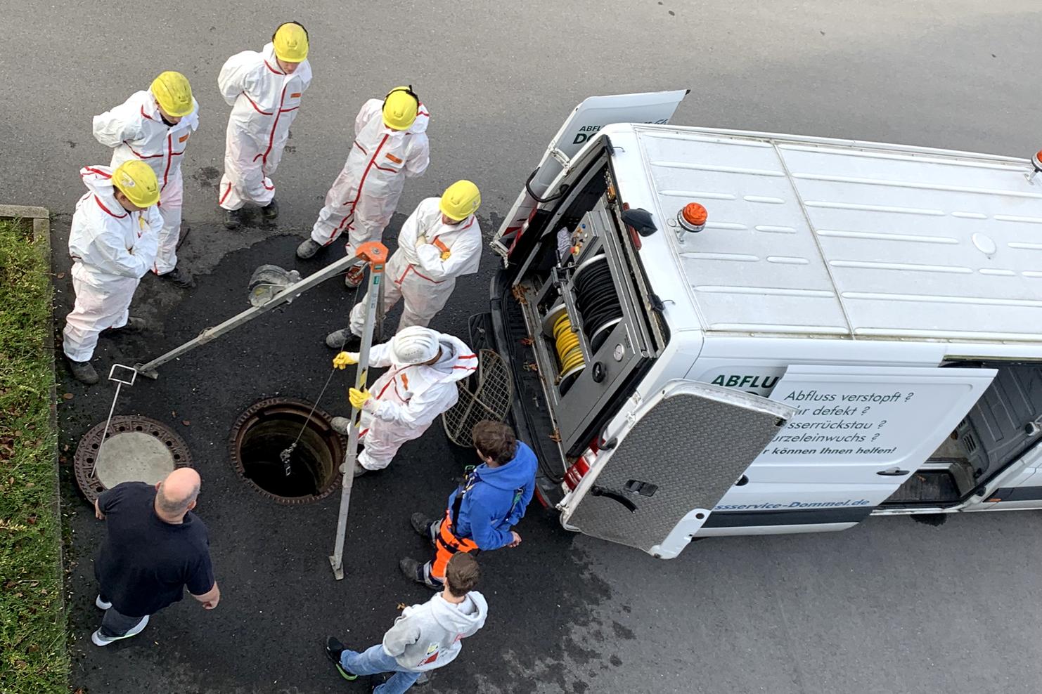 Gruppierung von sechs Fachkräften in Schutzanzügen am offenen Kanaldeckel, während ein Kollege mit Werkzeug arbeitet.