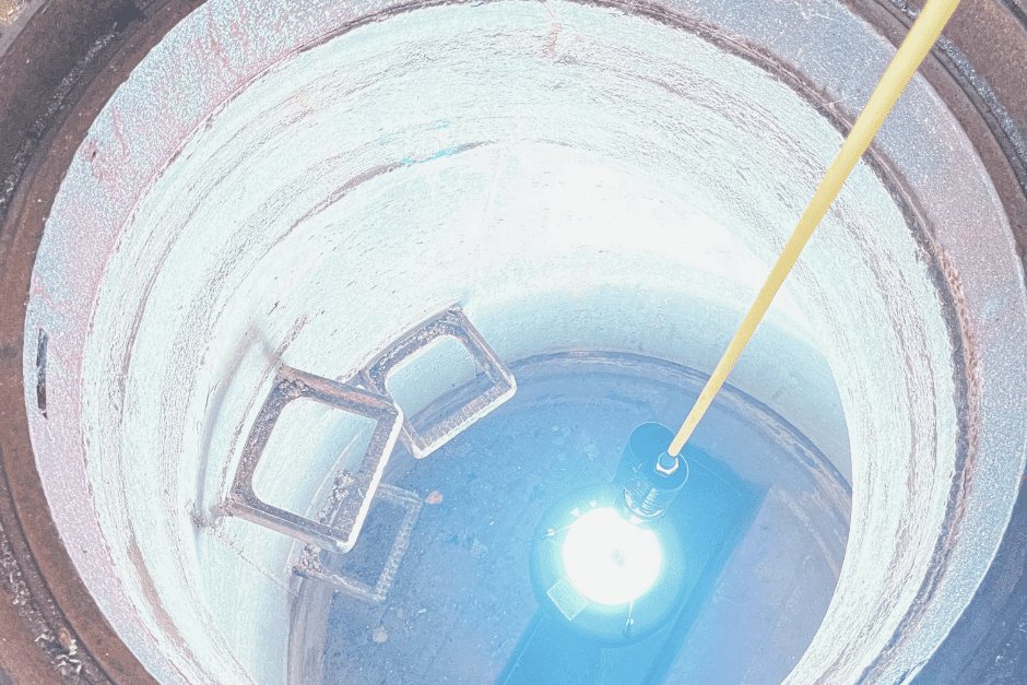 Blick in einen runden Schacht mit einer Lichtquelle und zwei rechteckigen Metallrahmen am Boden.