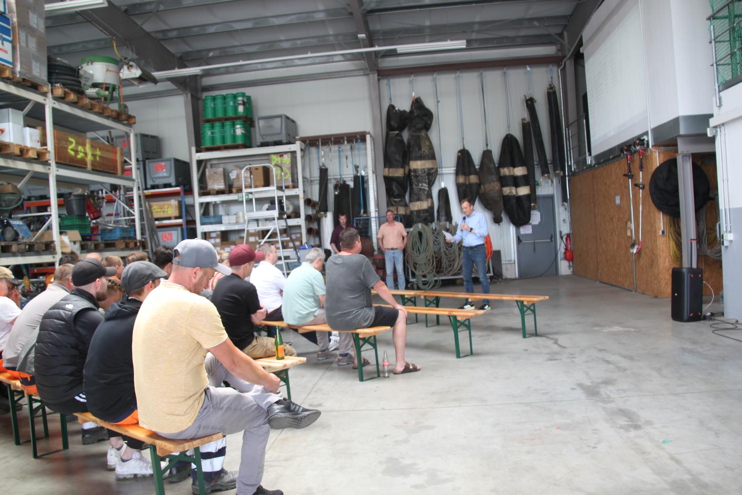 Gruppensitzung in einer Lagerhalle mit Teilnehmern, die auf Bänken sitzen, während ein Redner spricht.