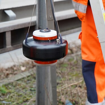 Gerät zur Vermessung hängt an einer Halterung neben einer Aluminiumleiter und einem Arbeiter in orangefarbener Kleidung.
