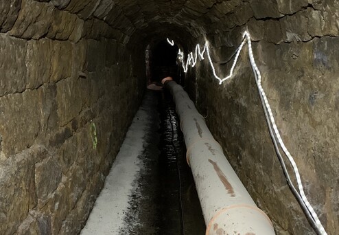 Ein schmaler, steinerner Tunnel mit einem großen, weißen Rohr und schwachem Licht am oberen Rand.