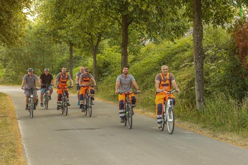 Sechs Männer in Arbeitskleidung radeln auf einem gepflasterten Weg unter Bäumen.