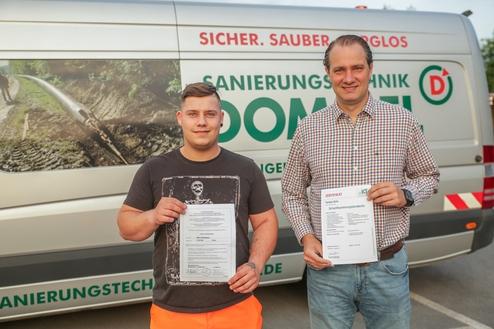 Zwei Männer halten Zertifikate in den Händen, vor einem Firmenfahrzeug mit dem Schriftzug "Sanierungstechnik".
