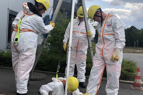 Vier Personen in Schutzanzügen und Helmen arbeiten an einem Schacht, eine Person ist teilweise darin.
