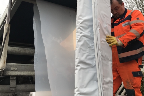 Arbeiter in orangefarbener Arbeitskleidung beim Entrollen einer weißen Plane von einem Lastwagen.